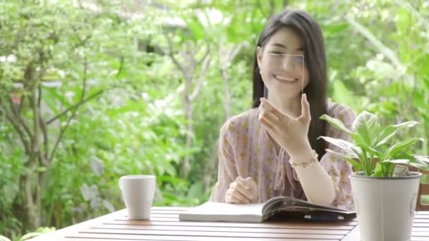 若い美しいアジアの女性のビデオは背景と笑顔で緑の木と彼女の家の庭でハイテクガラスディスプレイを使用して人に呼び出します 将来の技術コンセプト グラフィックオーバーレイの場合 — ストック動画