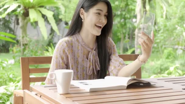 若い美しいアジアの女性のビデオは 背景に緑の木と彼女の家の庭でハイテクガラスディスプレイを使用して彼女の友人を呼び出し 笑っています 将来の技術コンセプト グラフィックオーバーレイの場合 — ストック動画