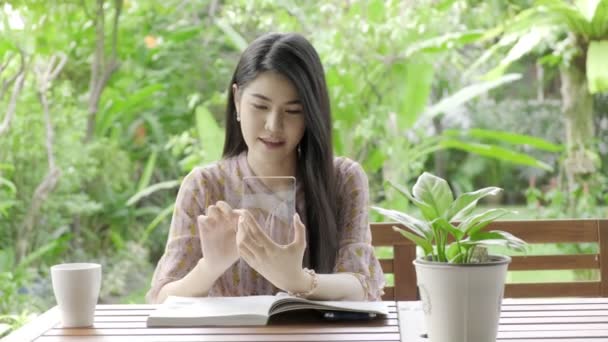 緑の木を背景に 幸せな笑顔で自宅の庭に透明ディスプレイを入力して若い美しいアジアの女性 将来の技術コンセプト グラフィックオーバーレイの場合 — ストック動画