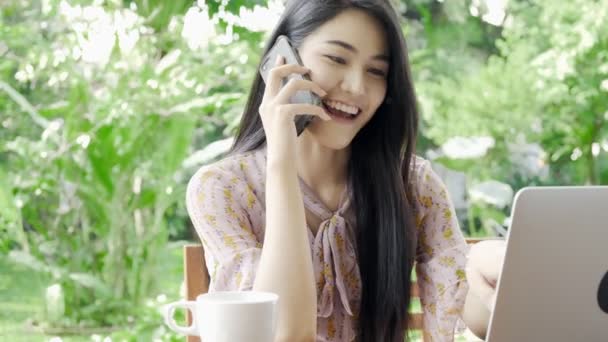 年轻迷人的亚洲女人通过电话与她的顾客交谈 并看着她的笔记本电脑在她家花园的绿树成荫 千年商业妇女生活方式概念 — 图库视频影像