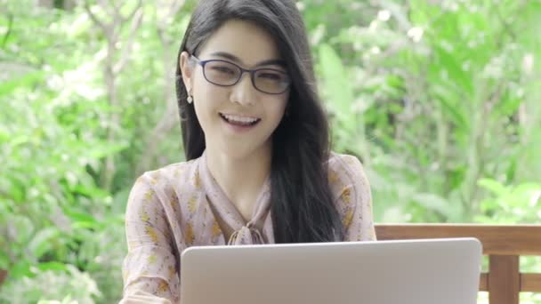 背景に緑の木と彼女の家の庭でラップトップとメガネでカメラを見て若い美しいアジアの女性のスーパースローモーション 幸せな笑顔 ミレニアルライフスタイルコンセプト — ストック動画