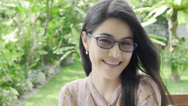 超级慢动作的年轻美丽的千禧年亚洲妇女戴着眼镜看着和微笑的相机在她的家庭花园与绿色树的背景 特写镜头 千禧年生活方式概念 — 图库视频影像