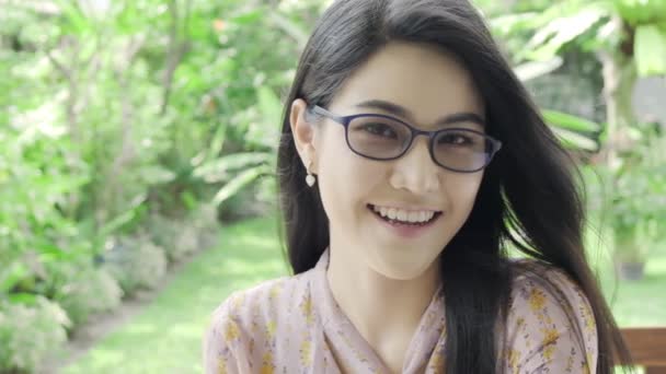 超级慢动作的年轻美丽的千禧年亚洲妇女戴着眼镜看着和微笑的相机在她的家庭花园与绿色树的背景 特写镜头 千禧年生活方式概念 — 图库视频影像