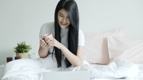 若い美しいアジアの女性がベッドでコーヒーを飲み ノートパソコンを使用して座っている 若いフリーランスのライフスタイル — ストック動画
