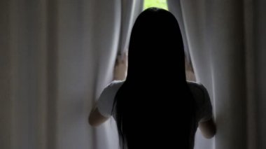 Genç ve güzel Asyalı kadın yeşil ağaçların arka planlarını görmek için yatak odasının perdesini açıyor. Rahatlatıcı ev kavramı.