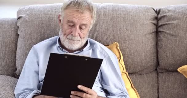 古いアジアの男性は 自宅でクリップボードに何かを書いています — ストック動画