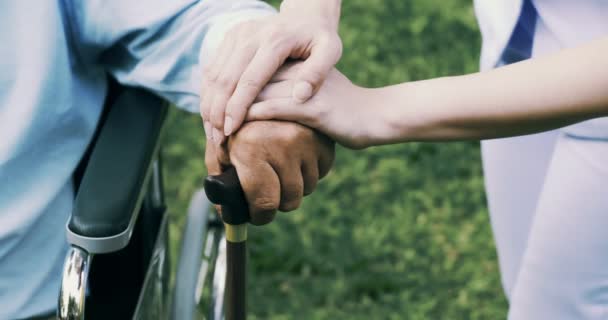 車椅子の若い看護師と成熟した障害者の男性は 庭でお互いに手を握っています — ストック動画
