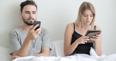 Çift yatakta cep telefonu ve tablet kullanıyor, ruh haline odaklanıyor. Beyaz erkek ve kadın..