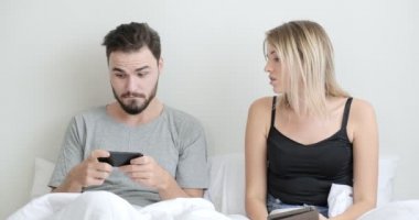 Yatakta cep telefonu ve tablet kullanan bir çift, bir kadın bir erkekten telefon alıyor, üzgün. Beyaz erkek ve kadın..