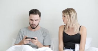 Yatakta cep telefonu ve tablet kullanan bir çift, bir kadın bir erkekten telefon alıyor, üzgün. Beyaz erkek ve kadın..
