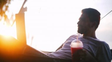 Günbatımında bir kokteyl onun elinde bir hamakta yatan bir dizüstü bilgisayar üzerinde çalışan genç, yakışıklı bir adam. Batan güneşin ışınları tropik bir adada tatile bilgisayarında çalışan genç iş adamı. Bir kokteyl yudumlarken ve çalışma serbest meslek 