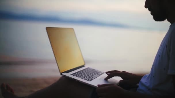 在落日下 这个年轻英俊的男人在海滩上的笔记本电脑上工作 日落时 年轻的商人在一个热带岛屿上工作 一个自由职业者在海边工作 — 图库视频影像