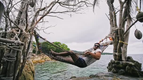 サングラスの男は 海沿いの熱帯の島の海岸にハンモックに横たわっています 海で休んでいる観光 — ストック動画