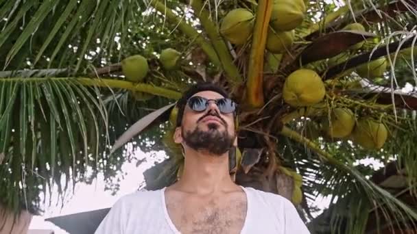 一个戴着太阳镜的英俊帅哥若有所思地向前看 那个穿白色 T恤的家伙站在一棵棕榈树下 — 图库视频影像