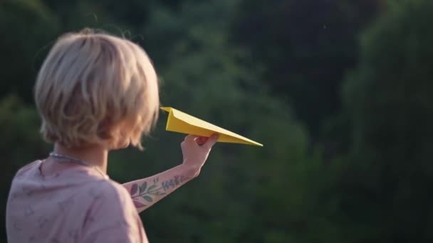Une jeune et belle fille tatouée à la main lance un avion en papier. Jolie femme, en plein air dans la nature, au coucher du soleil, lance un avion en papier coloré .