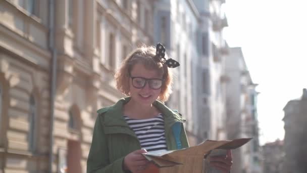 带着纸质地图的游客正在老街上寻找一条路 一个带着地图的年轻貌美的姑娘望着远方 戴眼镜的漂亮女人独自旅行 一个穿着绿色外套的可爱女孩走着笑着 阳光灿烂的日子 — 图库视频影像
