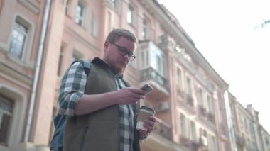 Sokakta kahve bardağı ve cep telefonu olan genç yakışıklı adam. Bir turist bir mobil cihaz üzerinde bir yol arıyor. Kablosuz teknoloji. Cep telefonundaki harita. Telefonda uygulama.