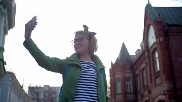 眼鏡をかけたフィンランド人の若い女の子と頭の上の弓は自撮りをします 美しい少女が旧市街で撮影されています 若い女性の観光客のアトラクションの背景に自分自身の写真を撮る 観光客が歩いて写真を撮る — ストック動画