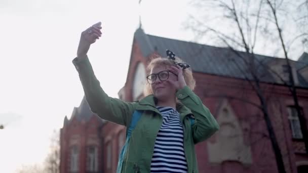 一个年轻漂亮的芬兰姑娘 戴着眼镜 头戴蝴蝶结 带着自拍 一个漂亮的女孩在古城被拍照 年轻的女游客在吸引人的背景下给自己拍照 一个游客散步和拍照 — 图库视频影像