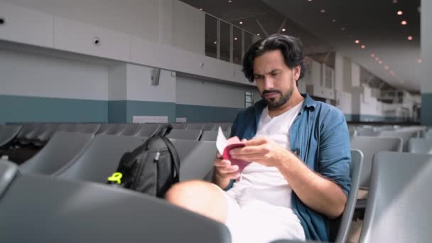 若いハンサムな男が待合室で一人で空港に座っている ハンサムな男がパスポートと切符を見ている フライトの前に空港で積極的な観光客 — ストック動画