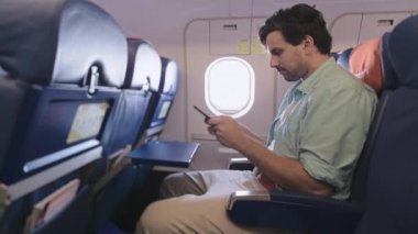 Çekici bir adam uçakta tek başına oturuyor ve elinde bir akıllı telefon tutuyor. Bir siteyi cep telefonuna kaydırmak. Sivil havacılıkta kablosuz teknolojiler. Uçakta kanepede bir turist oturuyor..