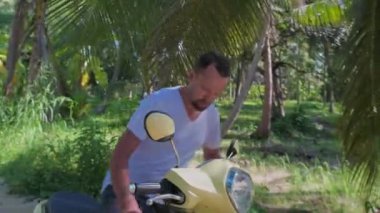 Genç, çekici bir adam sarı bir bisiklete oturuyor. Güneş gözlüklü ve beyaz tişörtlü yakışıklı bir adam yağmur ormanlarının arasında scooterını çalıştırıyor..