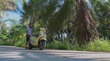 Genç, çekici bir adam sarı bir bisiklete oturuyor. Güneş gözlüklü ve beyaz tişörtlü yakışıklı bir adam yağmur ormanlarının arasında scooterını çalıştırıyor..