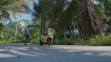 Genç, çekici bir adam sarı bir bisiklete oturuyor. Güneş gözlüklü ve beyaz tişörtlü yakışıklı bir adam yağmur ormanlarının arasında scooterını çalıştırıyor..