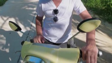 Genç, çekici bir adam sarı bir bisiklete oturuyor. Güneş gözlüklü ve beyaz tişörtlü yakışıklı bir adam yağmur ormanlarının arasında scooterını çalıştırıyor..