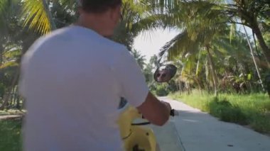 Genç, çekici bir adam sarı bir bisiklete oturuyor. Güneş gözlüklü ve beyaz tişörtlü yakışıklı bir adam yağmur ormanlarının arasında scooterını çalıştırıyor..