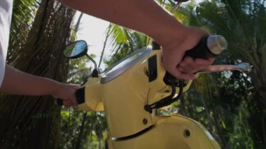 Genç, çekici bir adam sarı bir bisiklete oturuyor. Güneş gözlüklü ve beyaz tişörtlü yakışıklı bir adam yağmur ormanlarının arasında scooterını çalıştırıyor..