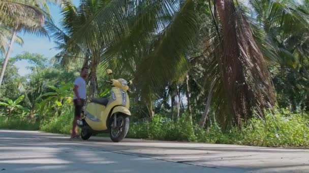 若い魅力的な男が黄色い自転車に腰を下ろしている 雨林の間の道路上でスクーターを起動します — ストック動画