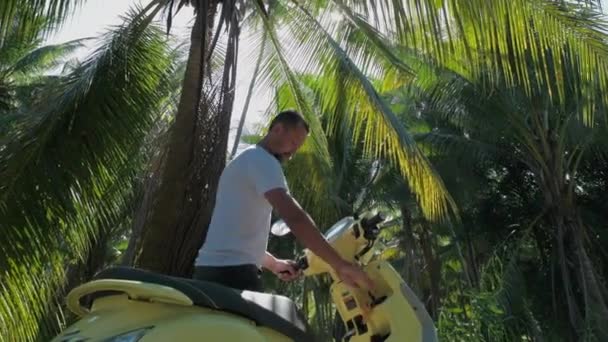 一个年轻貌美的男人坐在一辆黄色的自行车上 一个戴着太阳镜 身穿白色T恤的英俊男子在热带雨林的路上骑摩托车 — 图库视频影像