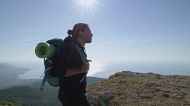 Genç, erkek bir turist dağlara tırmanıyor. Sırtında sırt çantası olan bir gezgin dağın tepesinde duruyor. Güneşin doğuşunda deniz manzarası.