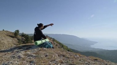 Genç bir turist yüksek bir dağdan deniz manzarasının tadını çıkarıyor. Bir adam, yükselen güneşin ışınlarıyla dağların tepesinde otururken termostan çay içer.
