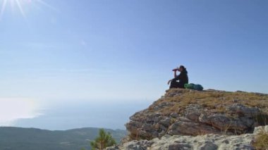 Genç, erkek bir turist dağlara tırmanıyor. Sırtında sırt çantası olan bir gezgin dağın tepesinde duruyor. Güneşin doğuşunda deniz manzarası.