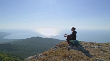 Genç bir turist yüksek bir dağdan deniz manzarasının tadını çıkarıyor. Bir adam, yükselen güneşin ışınlarıyla dağların tepesinde otururken termostan çay içer.