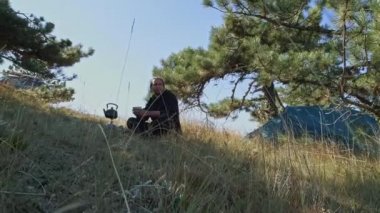 Kamp alanında genç ve yakışıklı bir adam gaz ocağını yakıyor. Kaynar su. Çadıra park etmek. Seyahat gaz ocağı. Bir turist fincandan çay içiyor.