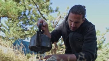 Kamp alanında genç ve yakışıklı bir adam gaz ocağını yakıyor. Kaynar su. Çadıra park etmek. Seyahat gaz ocağı. Bir turist fincandan çay içiyor.