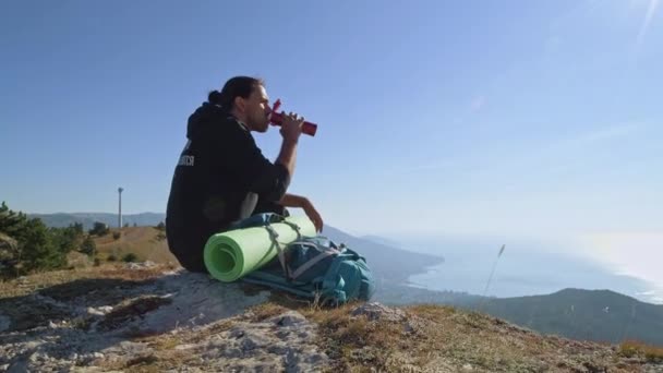 一位年轻的游客正在高山上欣赏海景 一个人坐在山坡上的悬崖上 沐浴在升起的阳光中 一边喝热水瓶里的茶 — 图库视频影像