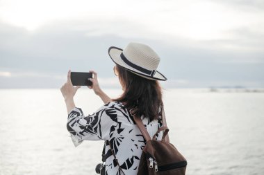 Gezgin genç kadının gündelik elbise holding smartphone ile plaj üzerinde tek başına duran sırt çantası içinde deniz arka plan vardır