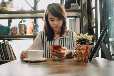 restoranda oturan ve cep telefonuyla genç kadın fincan kahve ve ahşap masa arka plan üzerine yerleştirilen bitkiler vardır