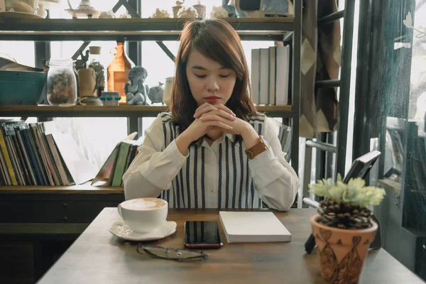 lifestyle background young woman sitting in restaurant and her eye close like thoughtful with cup of coffee, glasses, mobile phone and plant placed on wood table