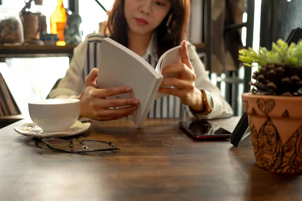 lifestyle background young woman sitting in restaurant and reading book with cup of coffee, glasses and plant placed on wood table