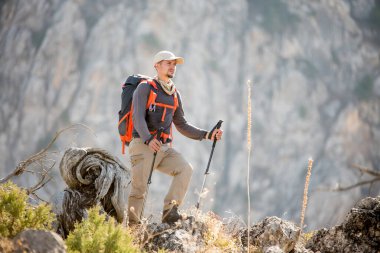 Turist kıyafetli bir adam bir dağın arka planında duruyor. Yazın doğa yürüyüşü, özgürlük gezisi. Kazanan üstte. Sırt çantası ve sırık taşıyan bir adam..