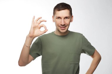 Neşeli mutlu beyaz erkek öğrenci geçirilen Sınav sonuçlarından memnun ok işareti yapar, rahat t gömlek giyer, yavaşça, beyaz arka plan üzerinde izole gülümsüyor. Tıraşsız adam hareketlerine kamera