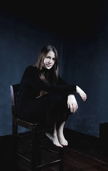 Cropped shot of attractive young stylish European female with healthy skin, looks seriously directly into camera, has natural beauty, poses on wooden chair, wearing black long dress and bare feet.