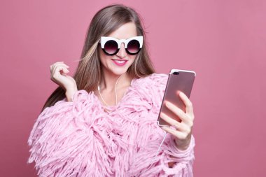 Güzel şık funky gür ceket giymiş, up olun giyer, akıllı telefon ve kulaklık üzerinden görüntülü görüşme yapar, olumlu ayakta pembe studio duvarın üzerinden, kamera gülümsüyor süre online arkadaşıyla konuşuyor.