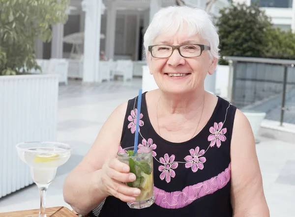 Adorable mature smiling relaxed grey haired female in dark blouse, enjoys spare time, recreation in cafe, tastes different cocktails, has happy expression. Relaxed woman tourist travels abroad.