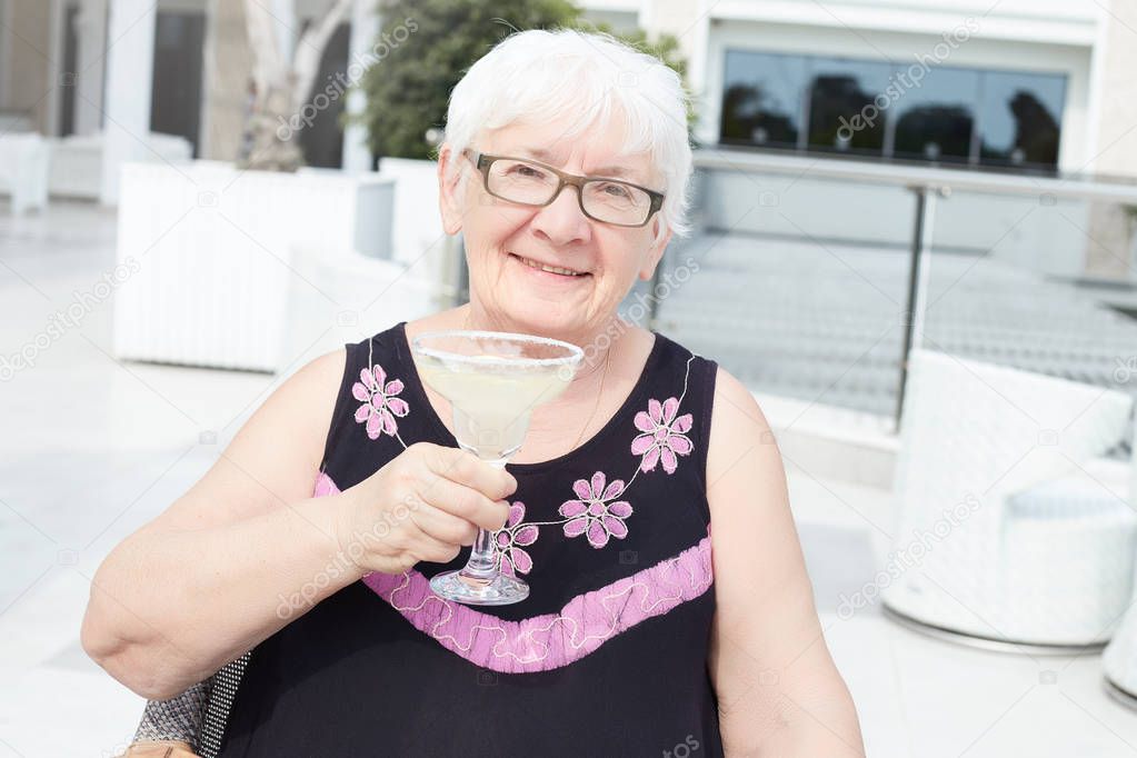 Lovely mature woman wears casual dress, drinks fresh cocktail, looks with dreamy expression at camera. Elderly female model with exotic beverage. Adorable adult woman recreates at open air cafeteria.